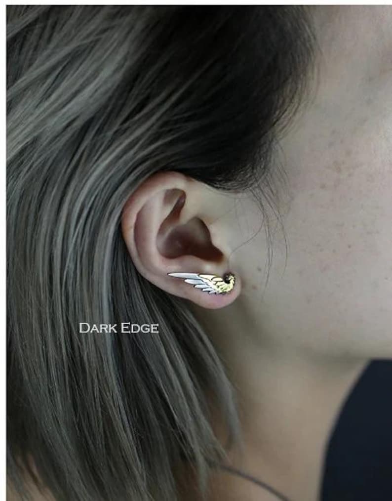 May include: A silver and gold wing earring with a delicate design. The earring is worn in the upper lobe of the ear.