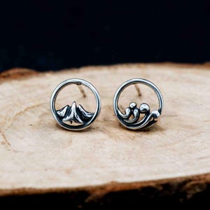 May include: A pair of silver stud earrings. One earring features a mountain design within a circular frame, while the other has a wave design. The earrings are set against a wooden surface.