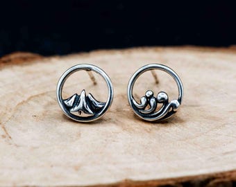 May include: A pair of silver stud earrings. One earring features a mountain design within a circular frame, while the other has a wave design. The earrings are set against a wooden surface.