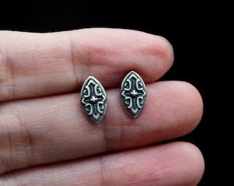 May include: A pair of silver-toned stud earrings. Each earring has an oval shape with a detailed, symmetrical design featuring a cross-like motif. The design is dark against the silver background, creating a contrast.