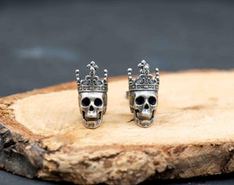 May include: A pair of silver-colored skull-shaped earrings, each adorned with a crown. The earrings are displayed on a natural wood surface, showcasing their intricate details and gothic aesthetic. The skulls have open mouths and detailed eye sockets.