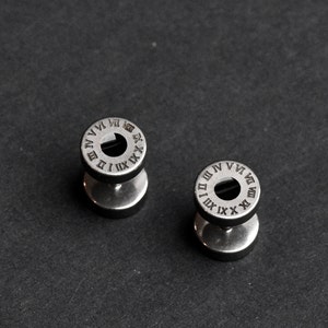 May include: Two silver-coloured metal ear gauges with a circular design featuring Roman numerals around the edge.