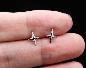 May include: A pair of small, silver-colored star-shaped stud earrings. Each earring has four points and a slightly rounded shape. The earrings are displayed against a dark background.