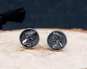 May include: Two silver-toned round stud earrings. Each earring features a profile of a woman with a crown in a circular frame. The earrings are displayed on a wooden surface with a dark blue background.
