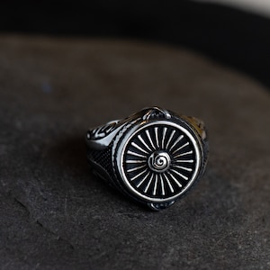 May include: A silver ring with a detailed design featuring a stylised jet engine. The ring has a black background and a silver spiral design in the centre.