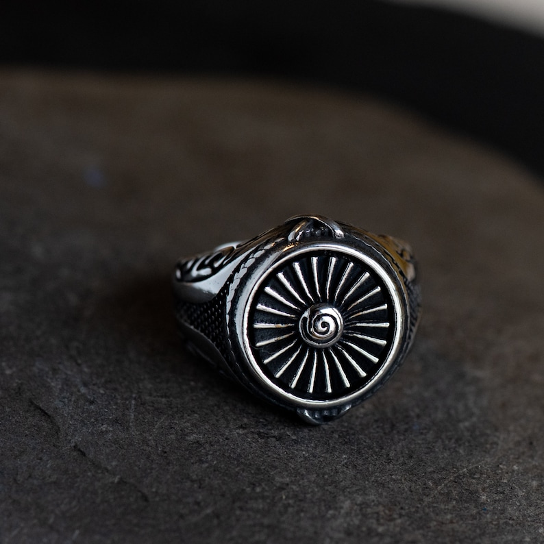 May include: A silver ring with a detailed design featuring a stylized jet engine. The ring has a black background and a silver spiral design in the center.