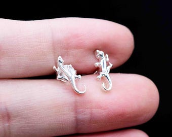 May include: A pair of small, silver-colored lizard stud earrings. The earrings are detailed with the lizard's body, legs, and curled tail. The earrings are displayed on a person's skin against a black background.