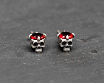 May include: A pair of silver-toned skull stud earrings. Each earring features a skull design with a red gemstone set in a crown-like setting. The earrings are displayed on a dark gray surface.