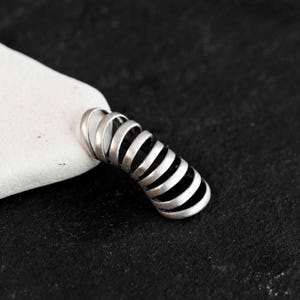 May include: A silver-colored ear cuff with a spiral design. The ear cuff is made of metal and has a series of curved bands that create a coiled effect. The cuff is resting on a white stone against a black background.