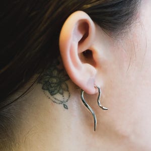 Op de afbeelding: Een zilverkleurige, gebogen oorbel wordt in het oor gedragen. De oorbel heeft een unieke, langwerpige vorm met een puntig uiteinde. Een bloementattoo is zichtbaar op de nek achter het oor.