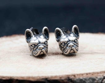 May include: A pair of silver-colored stud earrings. Each earring is shaped like a French Bulldog's head, with detailed facial features. The earrings are displayed on a wooden surface against a dark background. The design is detailed and the metal has a slightly aged appearance.