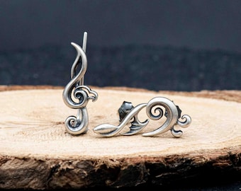 May include: A pair of silver-toned earrings in the shape of musical treble clefs. One earring is a stud, and the other is a climber. The earrings are displayed on a wooden surface with a dark background.