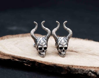 May include: A pair of silver-colored skull-shaped cufflinks with curved horns. The cufflinks are placed on a wooden surface against a dark background. The skull has detailed facial features.