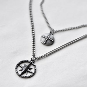 Pode incluir: Um colar de prata em camadas com dois pingentes. O pingente inferior é um design de rosa dos ventos dentro de uma moldura circular. O pingente superior é um pequeno pingente redondo com um design de cruz. As correntes são delicadas e os pingentes têm um acabamento antigo.
