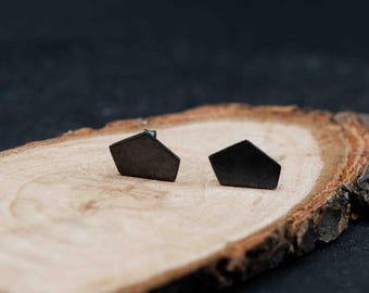 May include: Two dark gray, geometric stud earrings with a pentagon shape. The earrings are displayed on a natural wood slice with a dark background. The earrings are small and minimalist, suitable for everyday wear.
