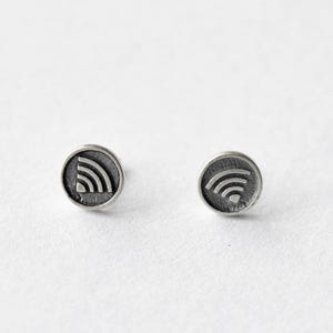 Könnte beinhalten: Ein Paar silberfarbene Ohrstecker. Jeder Ohrstecker hat ein kreisförmiges Design mit einem erhabenen WLAN-Symbol. Die Ohrringe sind vor einem weißen Hintergrund platziert, was ihren metallischen Glanz und ihr schlichtes Design hervorhebt.