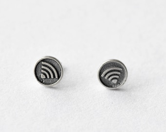 May include: A pair of silver-colored stud earrings. Each earring features a circular design with a raised Wi-Fi symbol. The earrings are set against a white background, highlighting their metallic sheen and simple design.
