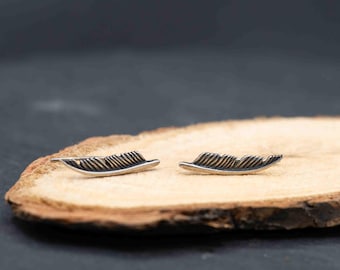 May include: A pair of silver-toned feather earrings. Each earring has a curved shape with detailed feather vein etching. The earrings are displayed on a light brown wooden surface against a dark background. The earrings are designed to be worn on the ear.