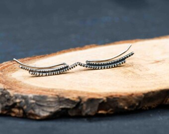 May include: A pair of silver-toned ear climbers with a curved design. Each earring features a series of small, evenly spaced rectangular details along the bottom edge. The earrings are displayed on a wooden surface.