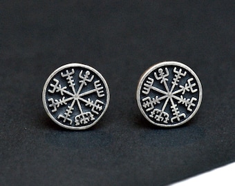 May include: A pair of silver-colored stud earrings. Each earring features a circular design with a central starburst pattern surrounded by stylized runes. The earrings are set against a dark background.