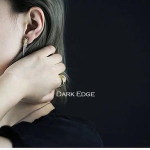 May include: A person wearing a silver earring with a gold accent and a gold ring on their finger. The text "DARK EDGE" is visible in the image.