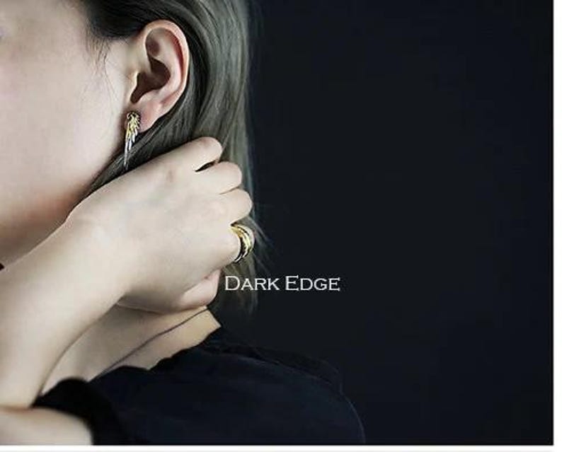 May include: A person wearing a silver earring with a gold accent and a gold ring on their finger. The text "DARK EDGE" is visible in the image.