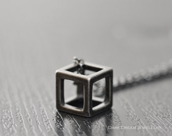 May include: A silver-toned pendant necklace featuring a hollow cube design. The cube is made of square metal bars, creating an open, geometric shape. The necklace has a delicate chain and is displayed on a dark, textured surface.