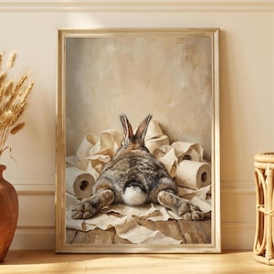 Puede incluir: Impresión enmarcada que representa un conejo descansando sobre rollos de papel. El conejo, visto por detrás, es marrón y blanco. La impresión está en un marco dorado. El fondo es beige neutro, con un jarrón y una pequeña mesa de mimbre.
