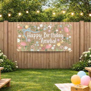 May include: A banner with the text "Happy Birthday Amelia!" is displayed on a wooden fence. The banner is decorated with floral and leaf designs in pastel colors. The scene is set outdoors with a wooden picnic table and a green lawn.