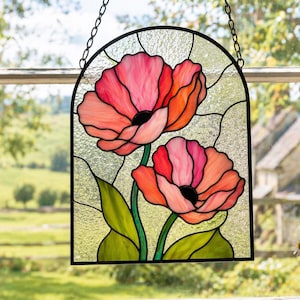 Könnte beinhalten: Buntglas-Fensterbild mit zwei leuchtend rosa und orangefarbenen Mohnblumen mit schwarzen Zentren und grünen Blättern. Das Panel ist schwarz umrandet und hängt an einer Kette. Der Hintergrund zeigt eine malerische Aussicht.