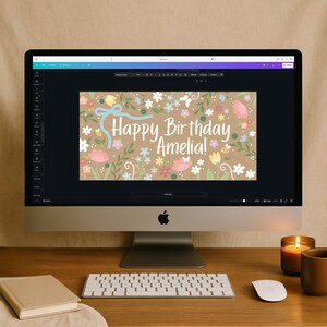May include: A desktop computer displays a birthday card design. The card features a floral pattern with pastel flowers, a blue ribbon, and the text "Happy Birthday Amelia!". A keyboard, mouse, book, candle, and mug are on the wooden desk.