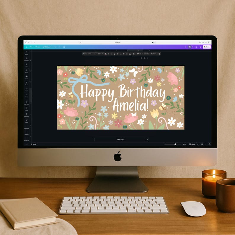 May include: A desktop computer displays a birthday card design. The card features a floral pattern with pastel flowers, a blue ribbon, and the text "Happy Birthday Amelia!". A keyboard, mouse, book, candle, and mug are on the wooden desk.