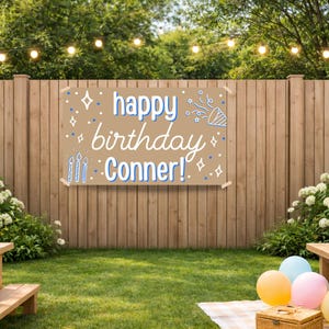 May include: A brown banner with the words "happy birthday Conner!" in white and blue lettering is displayed on a wooden fence. The scene is set outdoors with picnic tables, balloons, and string lights, suggesting a birthday party.