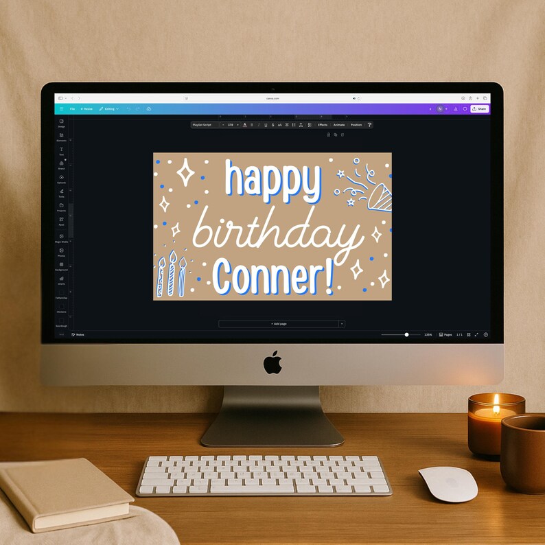 May include: A desktop computer displays a birthday card design with the text "happy birthday Conner!" in white and blue. The card features illustrations of candles, confetti, and stars. A keyboard, mouse, book, candle, and mug are on the wooden desk.
