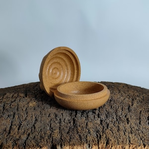 Può includere: Una piccola scatola rotonda in sughero con un coperchio. La scatola è aperta e l'interno è visibile. La scatola è realizzata in materiale di sughero naturale e ha un colore marrone chiaro.