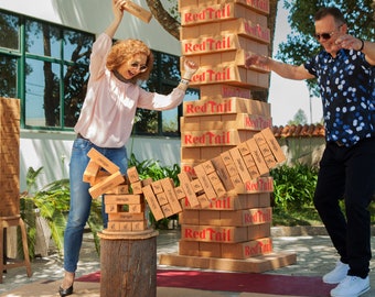 May include: Two people playing a game of giant Jenga with wooden blocks that have the words "Red Tail" printed on them. The blocks are stacked high and one person is about to pull a block out, while the other person watches.