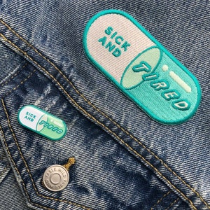 May include: A blue denim jacket with a white and green pill-shaped patch that says "Sick and Tired". The patch is sewn onto the jacket's pocket. There is a smaller, similar patch below it.