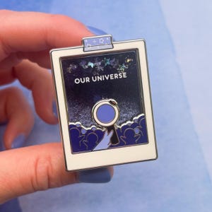 May include: A rectangular enamel pin with a white border and a dark blue background. The pin features the text "OUR UNIVERSE" and a design of a person looking through a magnifying glass at a crowd of people. The pin is held by a hand.