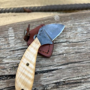 Könnte beinhalten: Ein kleines, handgefertigtes Messer mit einem hellen Holzgriff und einer gebogenen, silbernen Klinge. Der Griff hat zwei Messingnieten. Das Messer liegt auf einer Holzoberfläche mit einer braunen Lederscheide.