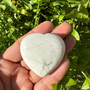 May include: A heart-shaped stone, light gray with subtle gray veining, is held in a hand. The stone's smooth surface contrasts with the rough texture of the hand. The background features green foliage.