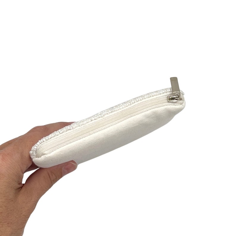 May include: A white zippered pouch with a beaded trim. The pouch is made of a soft, white fabric and has a silver zipper.
