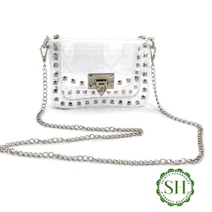 May include: A clear, rectangular crossbody bag with silver-tone hardware and a chain strap. The bag is adorned with silver studs and features a rectangular clasp closure. The bag is from Saints Headbands A Preppy Boutique.