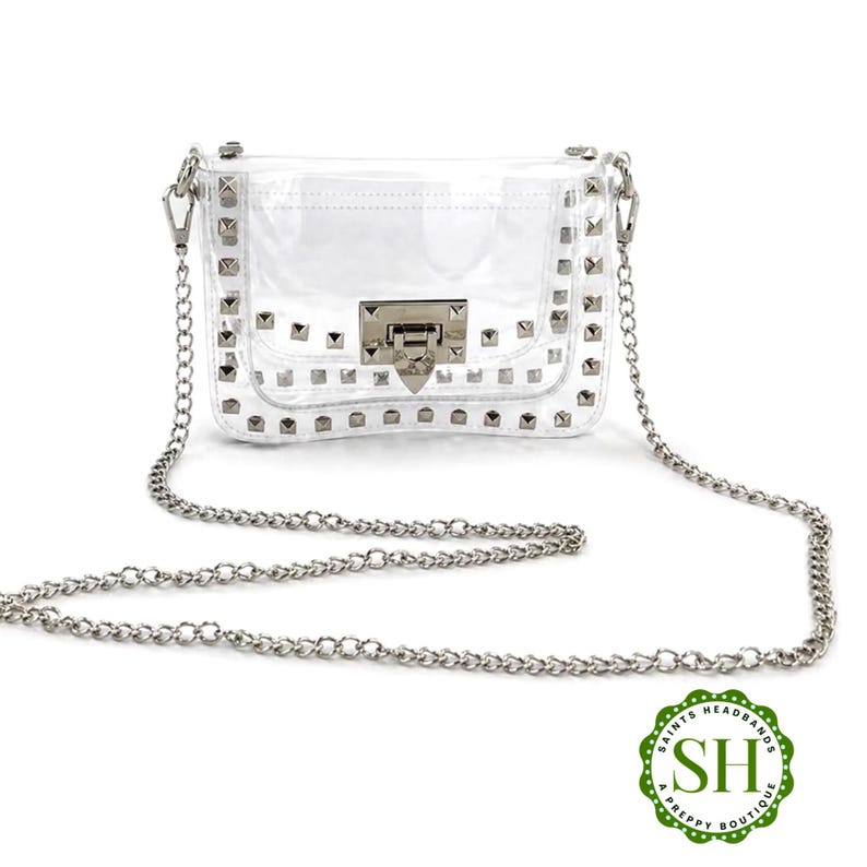 May include: A clear, rectangular crossbody bag with silver-tone hardware and a chain strap. The bag is adorned with silver studs and features a rectangular clasp closure. The bag is from Saints Headbands A Preppy Boutique.