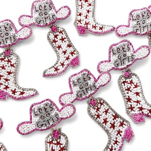 May include: A collection of beaded earrings shaped like cowboy hats and boots. The white hats feature the text "Let's Go Girls" in silver beads with pink trim. The white boots are decorated with red star patterns and pink heels, all crafted with beads.