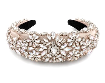 May include: A padded headband in a soft champagne satin fabric, densely embellished with clear, faceted crystals arranged in floral and marquise patterns. The interior of the headband is lined with a smooth black material for a secure fit.