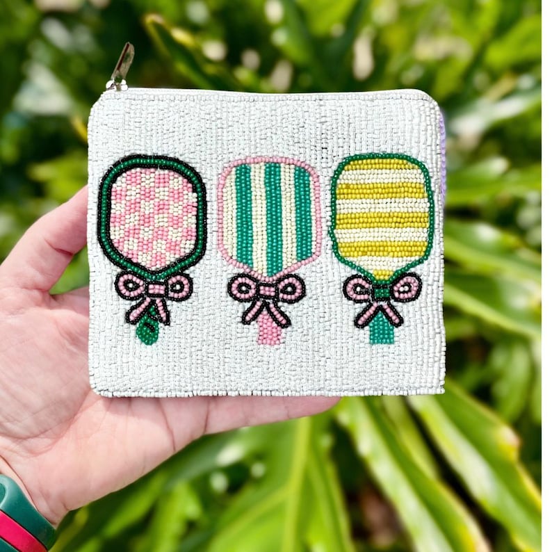May include: A white beaded zippered pouch with three beaded paddle designs. The paddles are pink and white checkered, green and white striped, and yellow and white striped. Each paddle has a pink bow.
