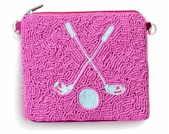 May include: A pink beaded square clutch with a zipper closure. The clutch features a design of two crossed golf clubs and a golf ball in light blue. The bag has a pink zipper and a small silver ring on each side.