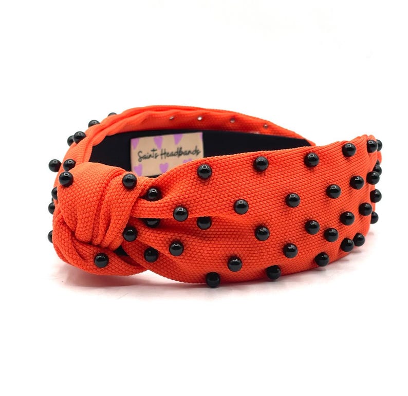 May include: Orange knotted headband with black beads. The headband has the text "Saints Headbands" on the side.