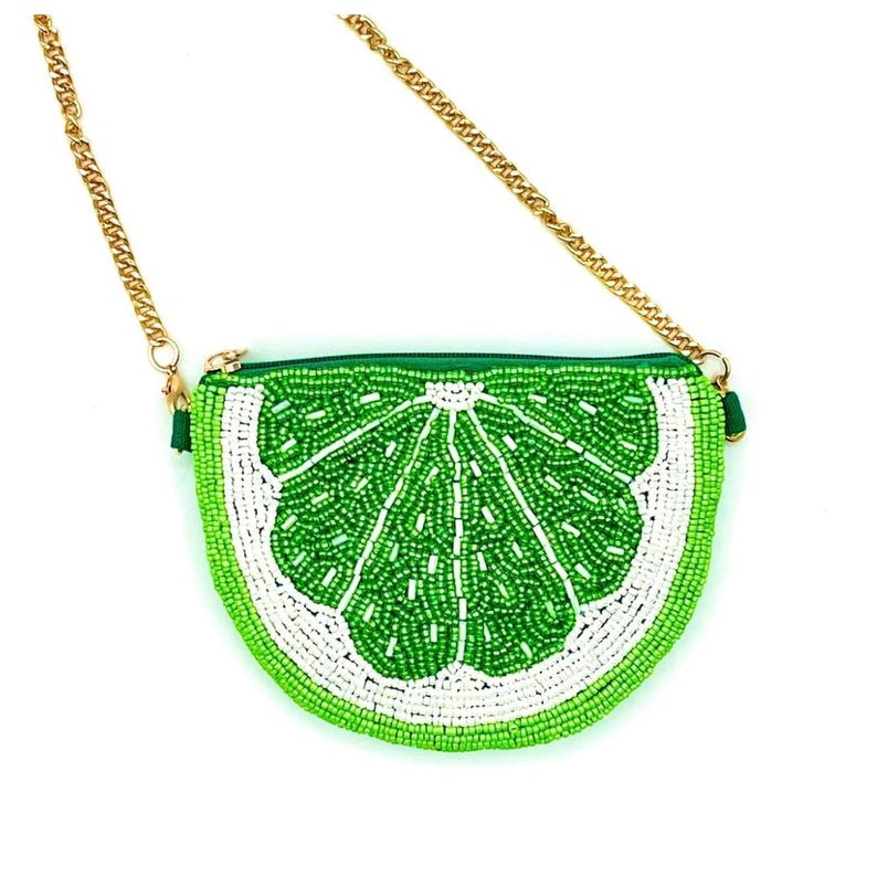 K&ouml;nnte beinhalten: Eine halbkreisf&ouml;rmige, perlenbesetzte Handtasche in Form einer Limettenscheibe mit leuchtend gr&uuml;nen und wei&szlig;en Glasperlen in einem Zitrussegmentmuster. Die Tasche verf&uuml;gt &uuml;ber einen goldfarbenen Metallkettenriemen, der mit gr&uuml;nen Schlaufen befestigt ist.