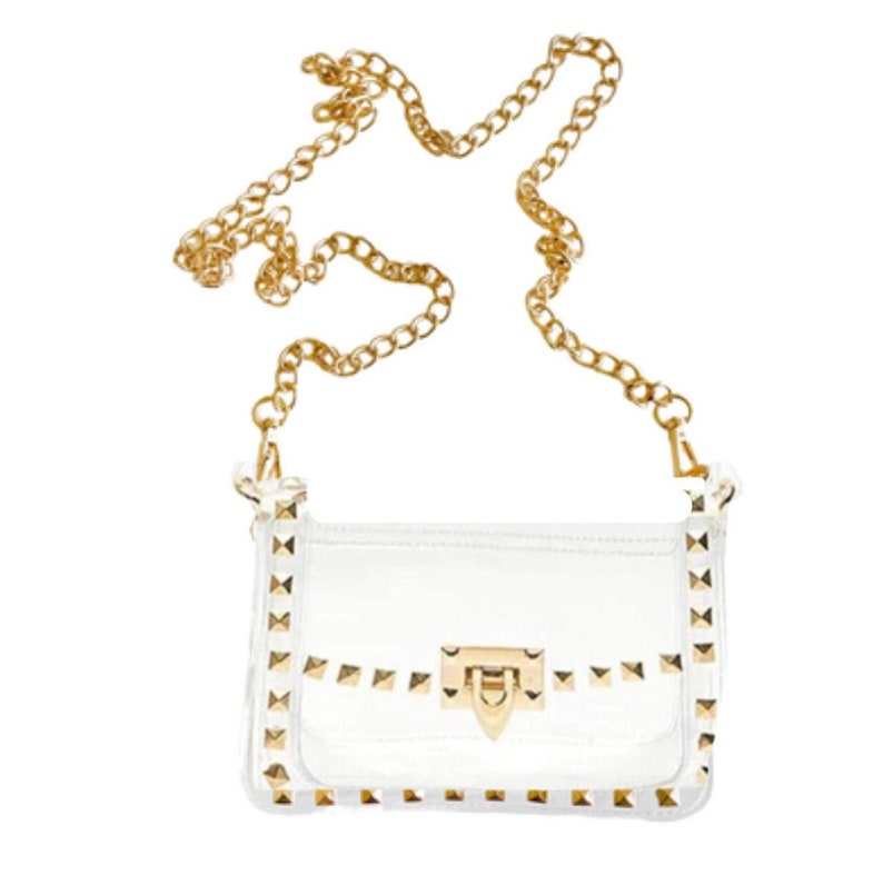 May include: A clear, rectangular crossbody bag with gold-colored chain strap and hardware. The bag is adorned with gold-colored pyramid studs along the edges and on the flap. The clasp is gold-colored.